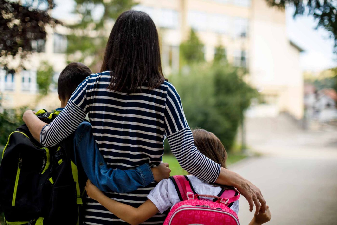 Woman with arms around children wearing backpacks walking down a sidewalk